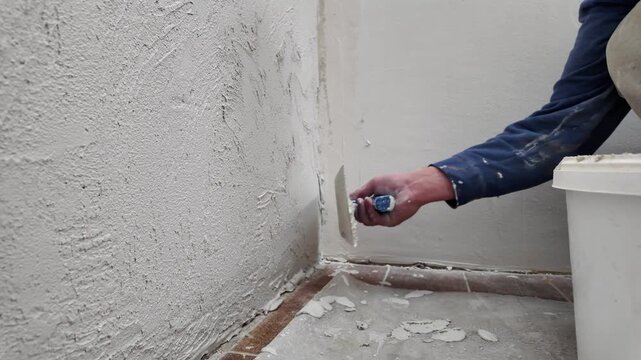 A worker prepares a wall by applying plaster to it. He uses a tool to spread the mixture evenly. The task takes place inside a home.