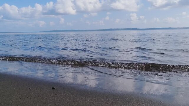 Waves gently roll onto sandy shore under bright blue sky with fluffy clouds