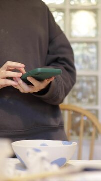 Unrecognizable female artist in a casual sweatshirt using a smartphone, texting and scrolling in her pottery studio, with a handmade ceramic bowl on the table
