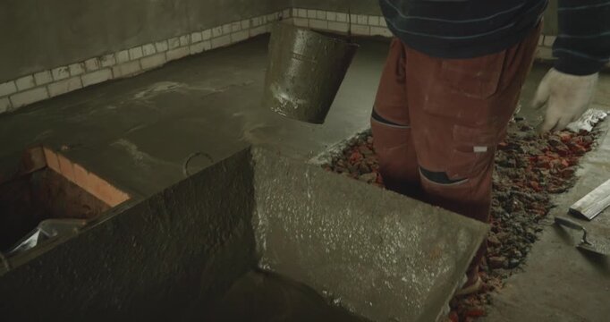 Male worker pouring wet cement mixture from a bucket to create a concrete floor screed at a construction site, preparing the foundation for a new building during renovation work