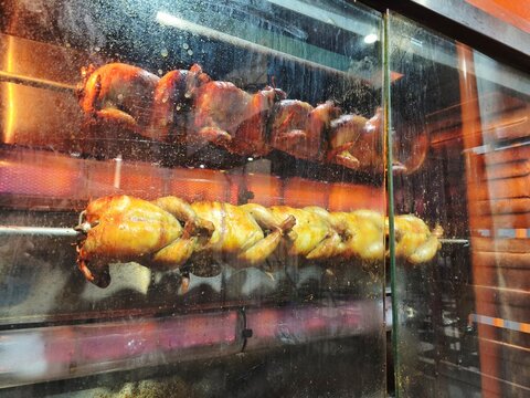 Rows of whole chickens being roasted in a rotating motion inside a modern rotisserie oven machine behind a restaurant glass window.