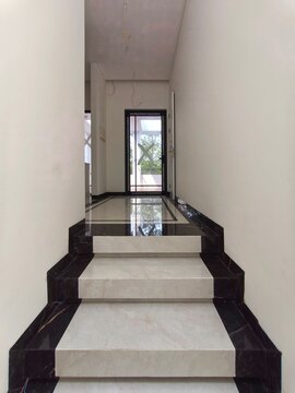 Modern minimalist house staircase interior design using white marble ceramic flooring with elegant glossy black granite border accents leading to a glass door.