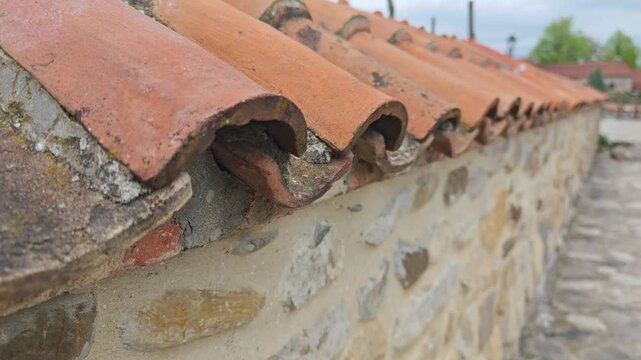 Close up of weathered terracotta roof tiles lining the top of an old stone wall. A reveal shot showing a rustic village street