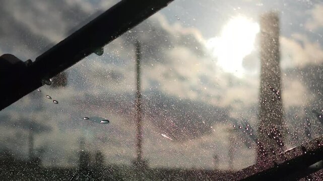 Car windshield wipers cleaning rain drops from the glass. View from inside the car on a rainy day with the sun shining through the clouds