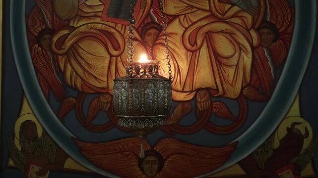 Solemn atmosphere inside an orthodox monastery with a swinging silver censer. The burning candle illuminates the religious mural of angels