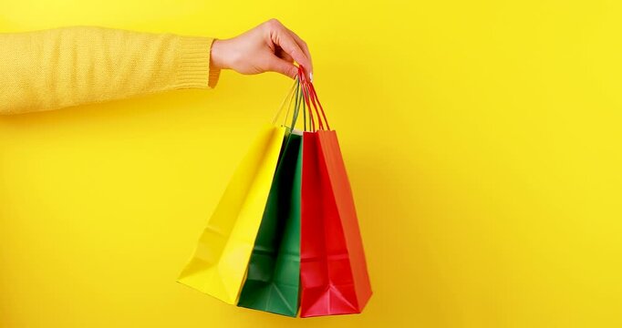 Close-up video of a hand holding and rotating three paper shopping bags in green, yellow, and red on a bright yellow background. Eco-friendly retail concept with reusable packaging and sustainable sho