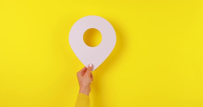 Close-up video of a hand holding a 3D location pin that appears and moves across the frame on a bright yellow background. Concept of GPS navigation, maps, tracking, travel, and geolocation services.