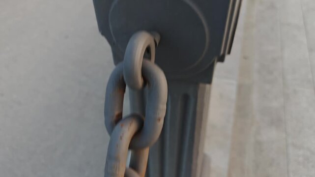 First person view moving along a thick, grey metal chain used as a barrier on a city sidewalk. The camera follows the rusty links