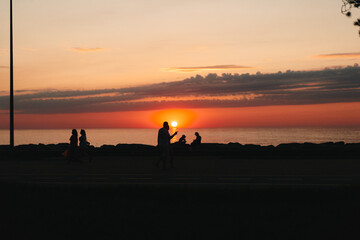Fototapeta premium Sunset beach scene with silhouettes of people, a calm coastal horizon, warm tones, a woman enjoying outdoor lifestyle, candid moment, gentle breeze, relaxed posture, vibrant sky