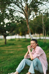 Fototapeta premium Cheerful woman outdoors, sitting on grass, smiling softly, relaxed posture, casual striped shirt, jeans, sunlit moment, gentle breeze, natural colors, serene park background
