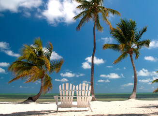 A double Adirondack chair on a sandy shoreline in Islamorada, Florida Keys, framed by palm trees and overlooking the still waters of the Atlantic Ocean under a bright afternoon sky © Michael