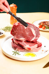 Fototapeta premium Chef cutting thick raw ribeye steak with knife on plate - fresh meat preparation in kitchen