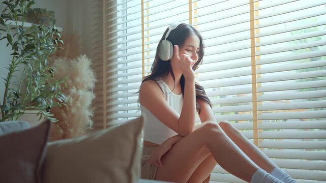 Happy Asian woman wearing headphones, singing and dancing while sitting by window at home, smiling young female listening to music or podcast in cozy bedroom for modern digital lifestyle and domestic
