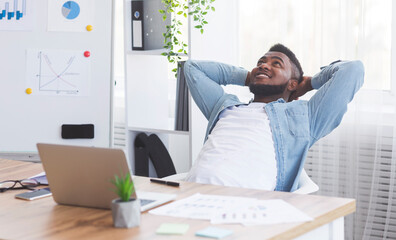 Obraz premium After productive day. Dreamy African American employee resting at workplace, sitting in comfortable chair with hands behind head and thinking about future.