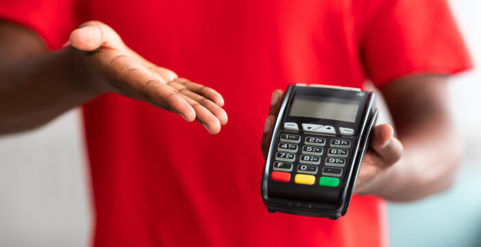 Digital Contactless Transaction. Cropped closeup view of African American man in red t-shirt uniform holding and showing POS machine, selective focus on payment dataphone terminal, blurred background