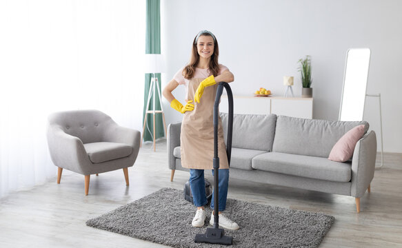 Housecleaning service. Happy young maid with vacuum cleaner standing in middle of tidy living room, full length portrait. Positive millennial housewife doing domestic chores, copy space