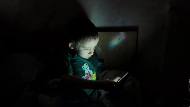 A child is sitting on a bed in a dark room, illuminated only by the light from a smartphone. The child is focused on the screen, unaware of the surrounding darkness. This moment captures the interacti