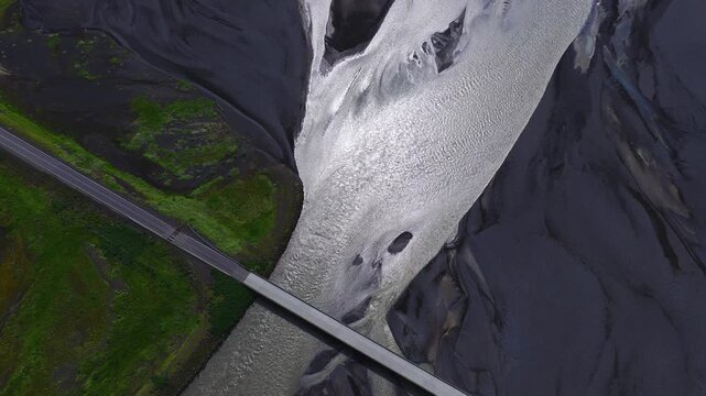Aerial view shows a straight bridge over braided glacial water near Seljalandsfoss in southern Iceland, with black sand, green moss, ripples, islands, and one car.