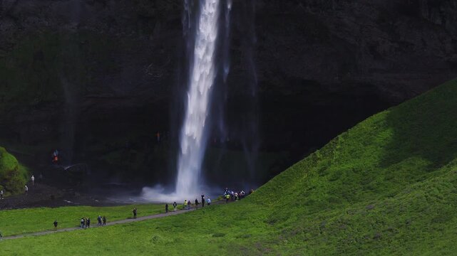 Aerial view of Seljalandsfoss in southern Iceland as water plunges from a basalt cliff into a misty pool. Tourists walk the footpath and the walk behind cavern shows.