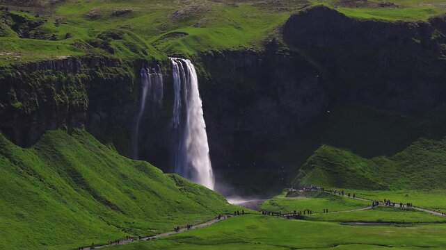 Aerial view of Seljalandsfoss waterfall in southern Iceland shows water plunging over a basalt cliff into mist, with riverside path, tiny people, and sunlit grassy ridges.