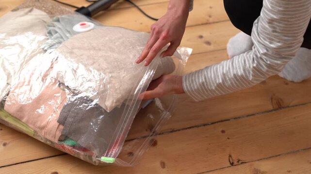 A woman neatly places folded clothes into a vacuum compression bag on a wooden floor. She carefully arranges the items and then seals the bag to save space for storage or travel
