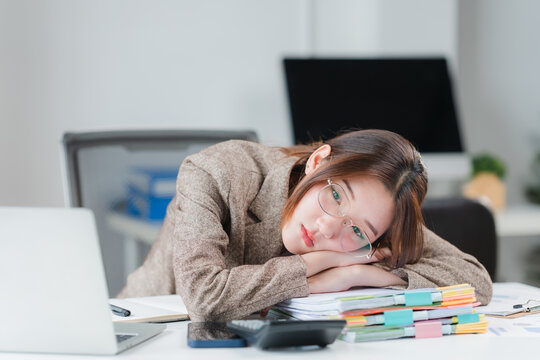Overworked Professional: A businesswoman rests her head on a stack of papers on her desk, appearing exhausted and overwhelmed, a common sight in demanding work environments.