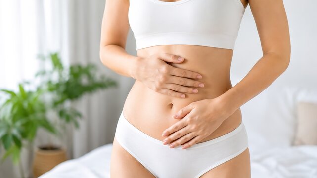 A young woman wearing a white crop top and panties is seated on a bed holding her stomach with both hands expressing discomfort or pain in a calm and peaceful bedroom setting