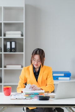 Focused Professional at Work: A woman in a vibrant orange blazer meticulously reviews documents at her desk, surrounded by office supplies, including a laptop and calculator, in a bright.