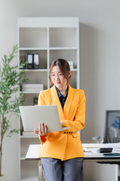 Young Woman with Laptop in Office: A focused young woman in a bright yellow blazer stands in a modern office, holding a laptop and looking thoughtfully at the screen.