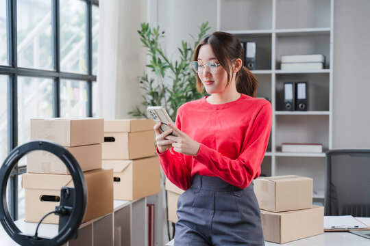 Online Seller at Work: A young Asian woman, engrossed in her smartphone, stands amidst a backdrop of neatly stacked cardboard boxes.
