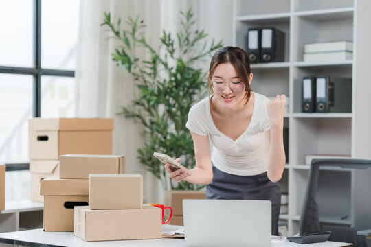 Small Business Success: A young Asian woman, an entrepreneur, celebrates a successful sale while working from home, surrounded by shipping boxes and her laptop.