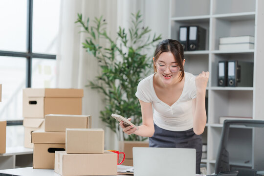 Entrepreneurial Triumph: A young Asian businesswoman, brimming with excitement, celebrates an online sale in her home office, surrounded by an array of shipping boxes and business supplies.