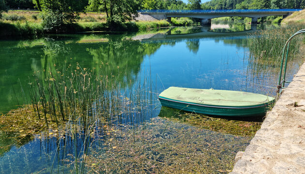 A peaceful scene on the Dobra river featuring a small green boat moored by a stone bank next to clear water and a distant road bridge.