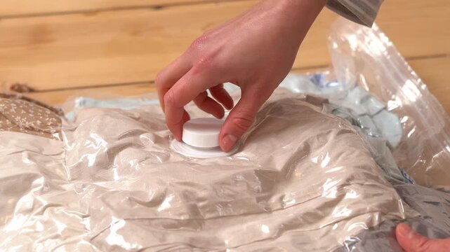 A person's hands twist the white cap shut on a vacuum storage bag full of clothes, set on a light wood floor