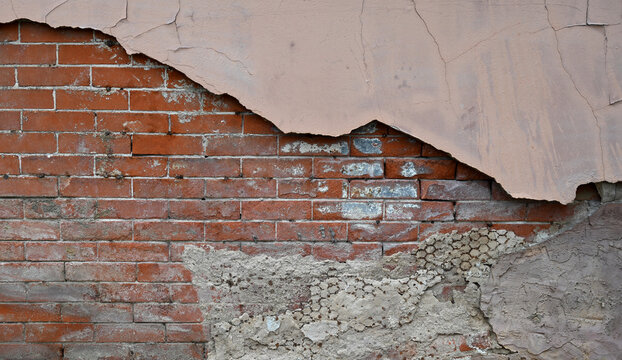 Horizontal close up of a weather beaten tan painted decaying wall surface with cracked plaster, exposed concrete and metal lath, rough cement layer and red brick with worn mortar joints