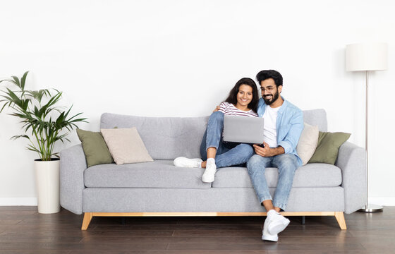 Loving happy beautiful eastern couple in stylish casual outfit sitting on couch at home, embracing, using laptop, young indian man and woman watching movie online, copy space, full length