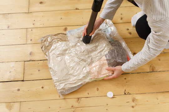 High angle view of a person using a vacuum to compress clothes in a storage bag on a wood floor. Home organization, decluttering, space saving concept for moving seasonal storage. Natural light