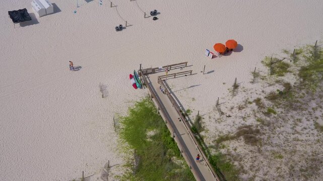 Aerial view of a wooden beach boardwalk leading to pristine white sand in Gulf Shores / Orange Beach, Alabama. People walk along the path toward bright orange umbrellas and the Gulf of Mexico beach.