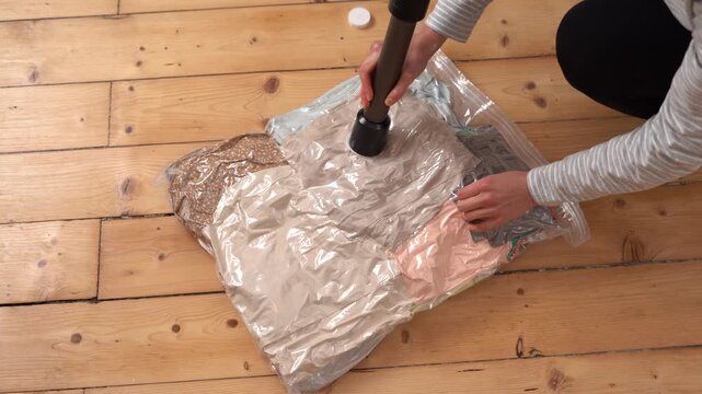 A woman uses a vacuum cleaner to remove air from a space saver bag full of clothes. The bag compresses, shrinking the garments for compact storage or travel packing. Filmed on a wooden floor
