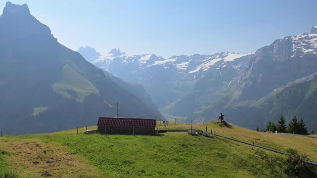 Summer mountain view with green meadow, rustic hut and distant snowy peaks along the Walenpfad hiking route above Engelberg, Switzerland.