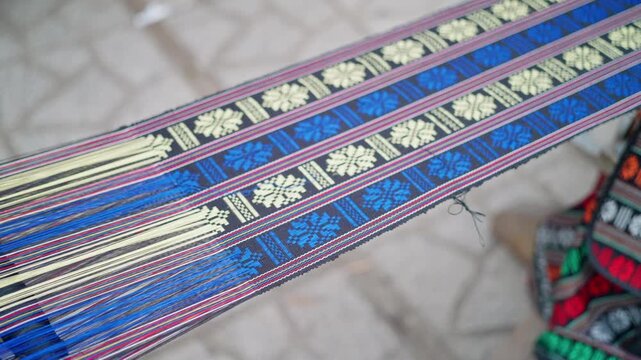Handwoven textile band on loom with taut multicolored warp threads and patterned blue cream ribbon, showing delicate crafting movement under soft diffused workshop light