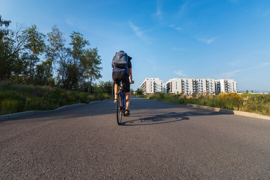 Back view of a man with a backpack riding a bicycle on an asphalt path heading towards a modern residential area