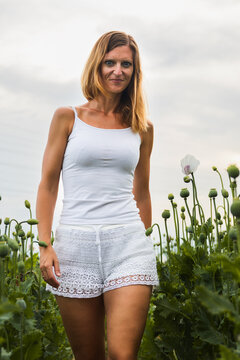 Portrait of a smiling woman in white clothes walking through a poppy field with green seed pods and white flowers