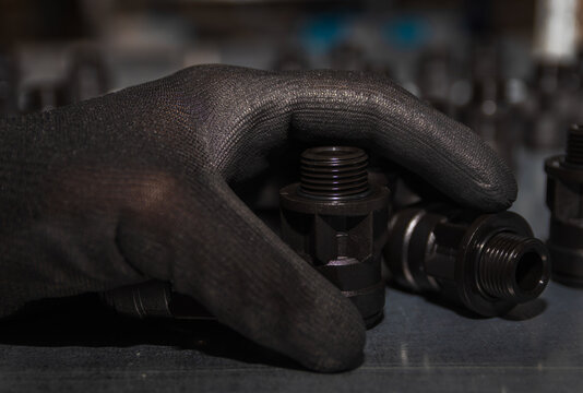 Close-up shot of a hand in a black protective glove holding black plastic threaded pipe fittings on a shelf in a workshop