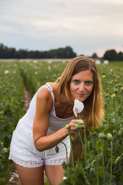 Portrait of a woman in white clothes leaning down and smelling a blooming white poppy flower in a large field