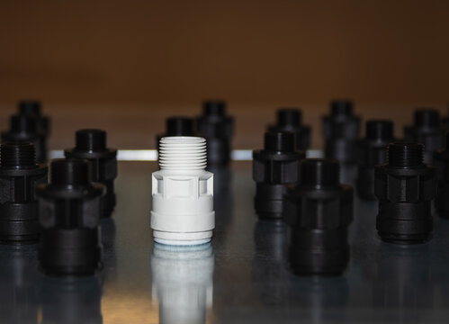 Front view from a low angle of a single white plastic pipe fitting standing in the foreground ahead of rows of black connectors