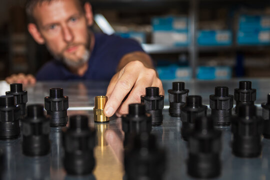Warehouse worker placing a single brass threaded pipe fitting among rows of black plastic connectors on a metal table