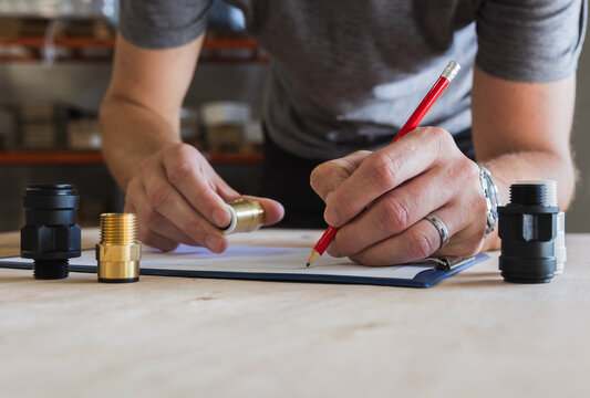 Close-up of a worker writing with a red pencil on a clipboard checklist during quality control of pipe fittings