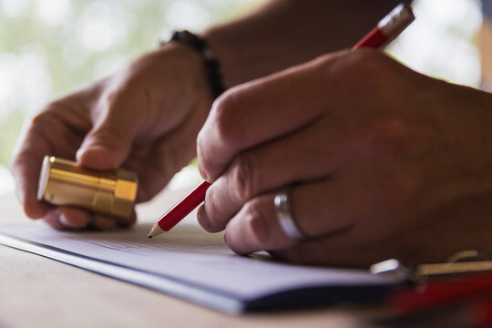 Extreme close-up of technician hands writing on a clipboard with a red pencil while holding a brass component