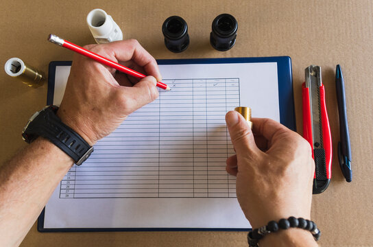 Flat lay top view of inspector hands writing with a red pencil on a clipboard checklist during brass and plastic pipe fittings inspection